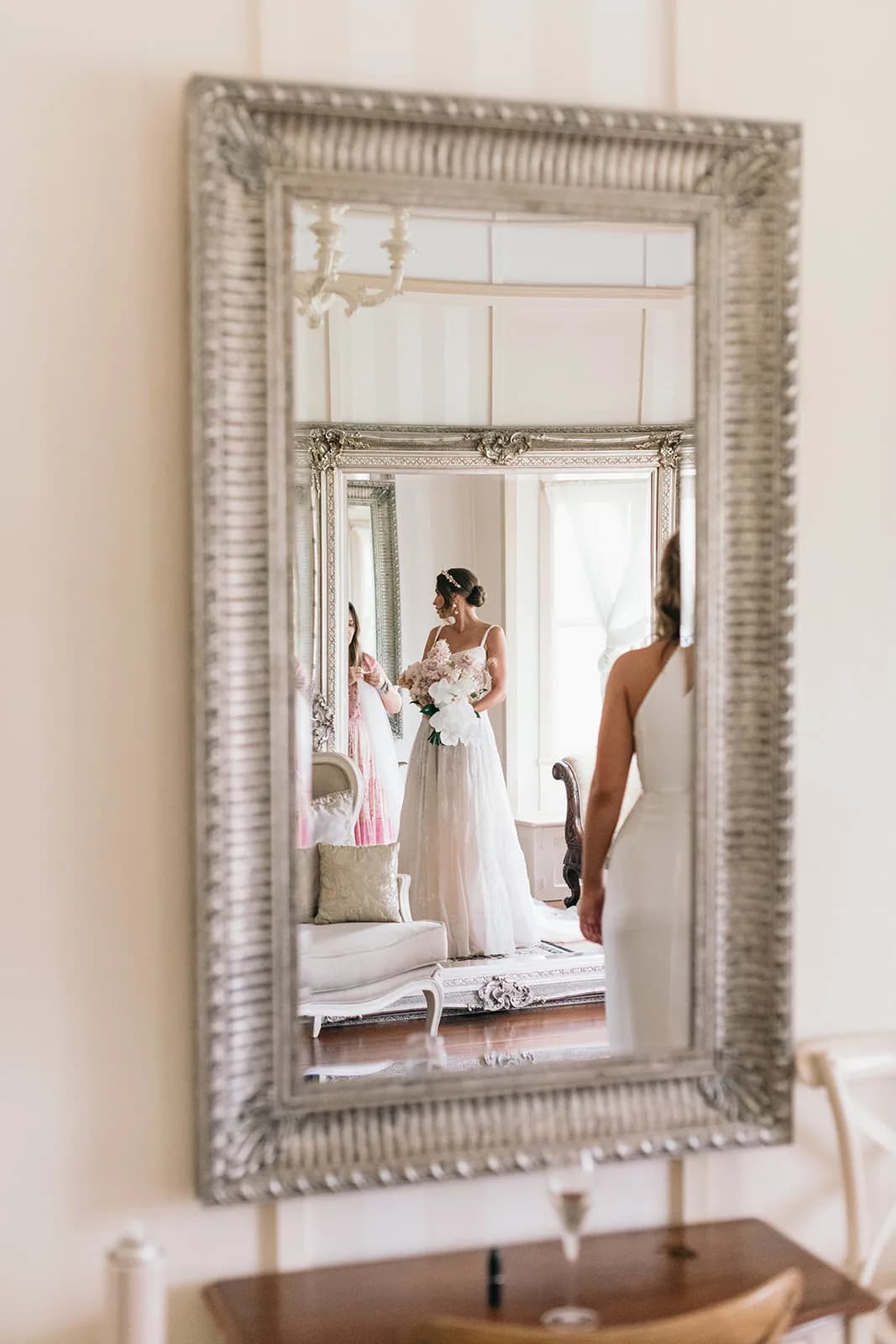 A bride stands in front of a large ornate mirror, holding a bouquet of white flowers and looking at herself. A bridesmaid in a white dress stands nearby. The scene is reflected in the mirror, with elegant furniture and soft lighting in the background.