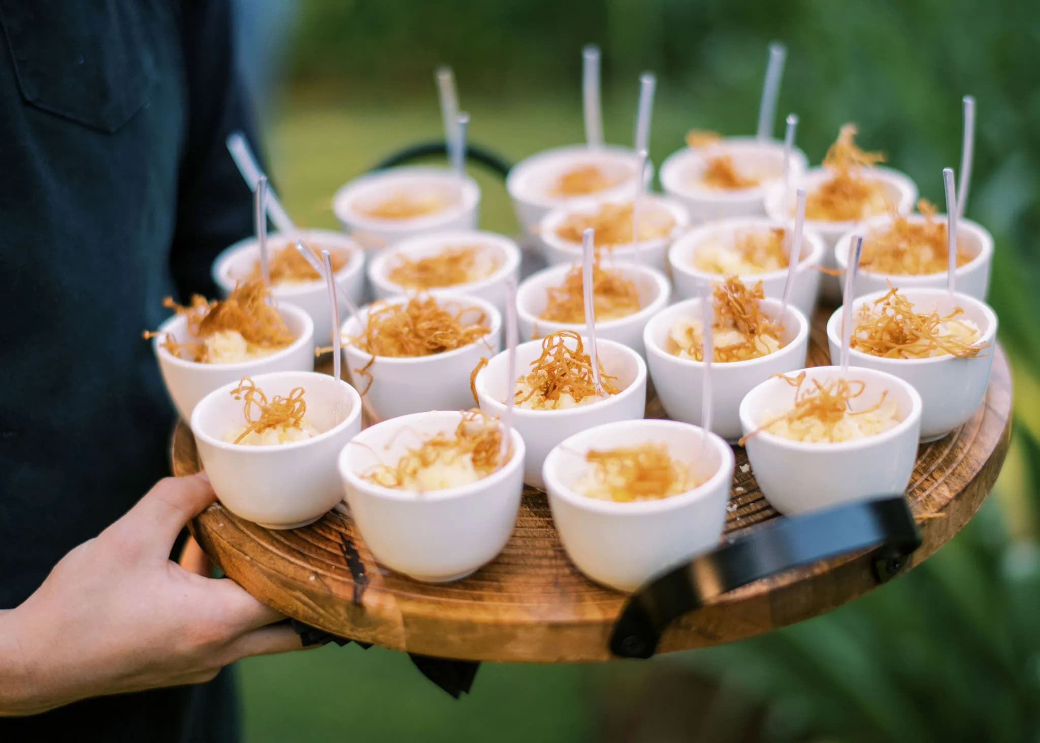 A person holds a round wooden tray filled with small white bowls of a dish topped with thin, crispy, fried strands and metal spoons. The background is blurred, suggesting an outdoor setting.