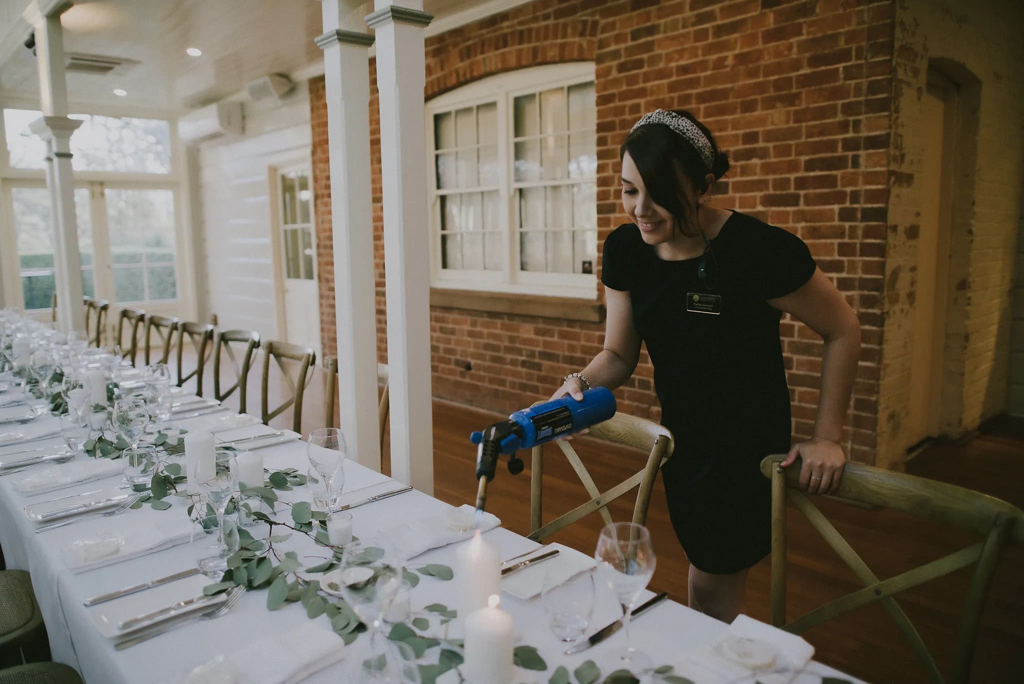 A woman in a black dress is lighting candles using a blue device along a long, elegantly set dining table. The table is adorned with greenery and white linens, set with glassware and cutlery. The setting is inside a rustic venue with exposed brick walls.