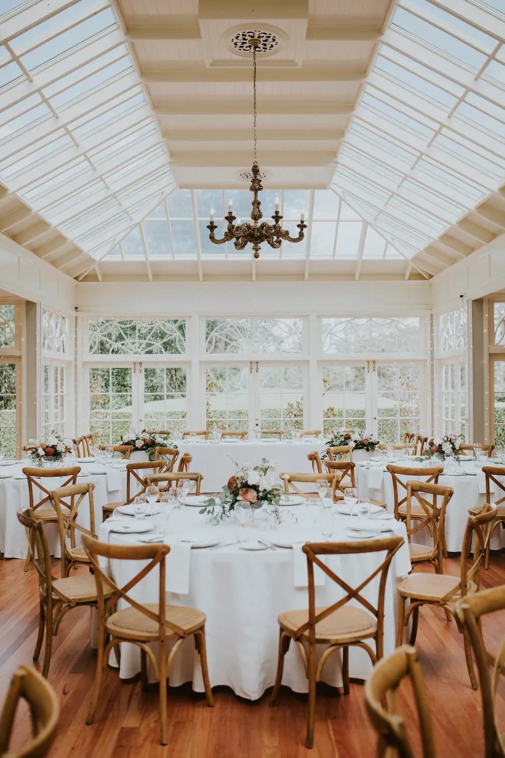 An elegant sunlit reception hall with a high glass ceiling and chandelier. Round tables with white tablecloths and floral centerpieces are arranged neatly. Wooden chairs surround the tables, and large windows offer a view of greenery outside.
