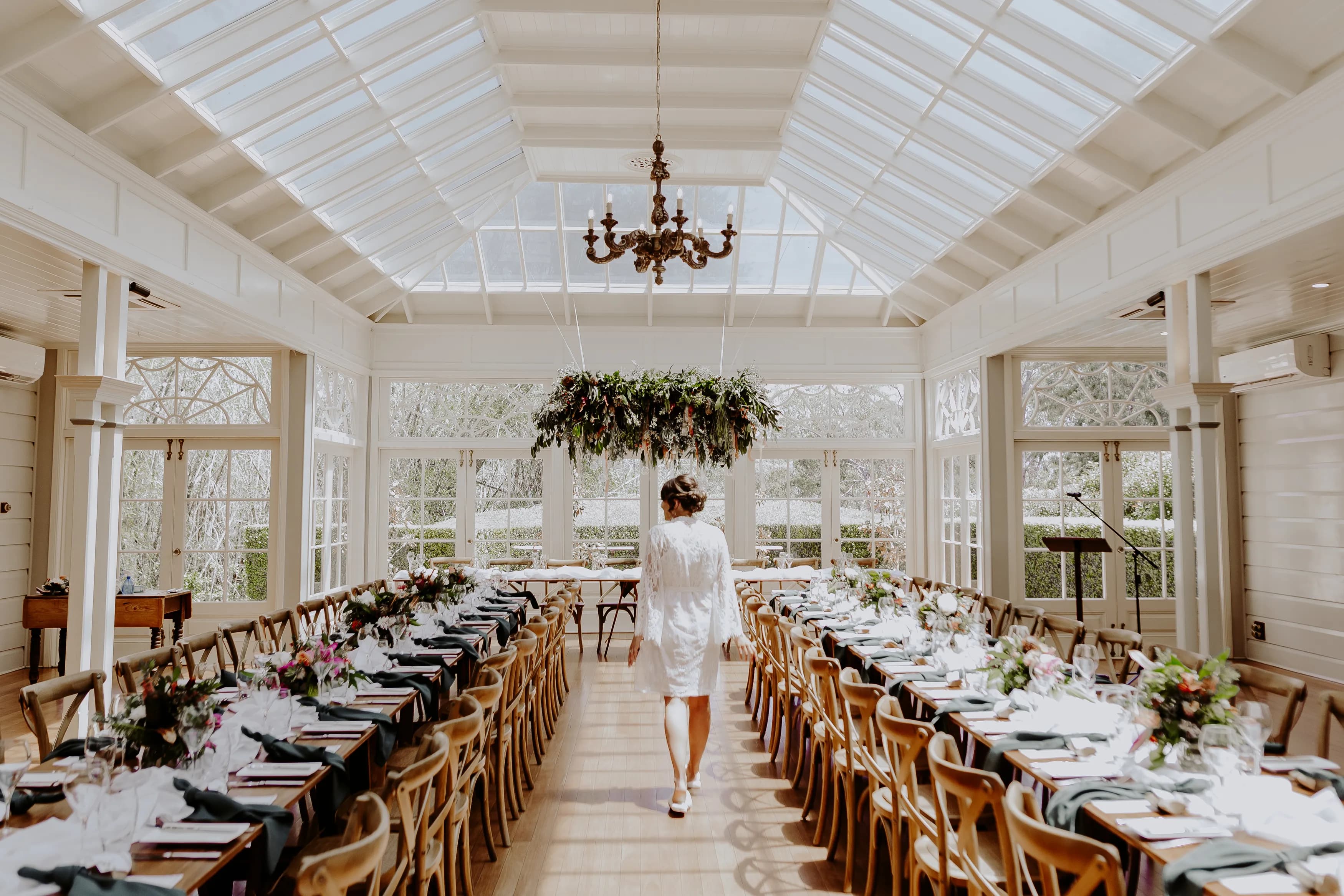 A person in white attire stands in the center of a beautifully decorated banquet hall with large glass windows and a skylight ceiling. Long tables, adorned with floral arrangements and place settings, line both sides of the room. A hanging chandelier is visible overhead.