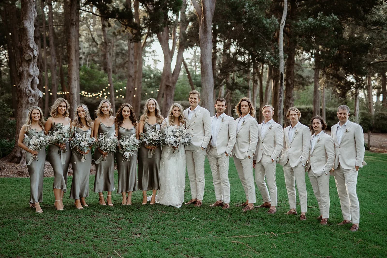 A wedding party stands outdoors on green grass with a backdrop of trees and string lights. The bridesmaids wear matching silver dresses, and the groomsmen wear light beige suits. The bride, in a white gown, and the groom, in a matching suit, stand in the center.