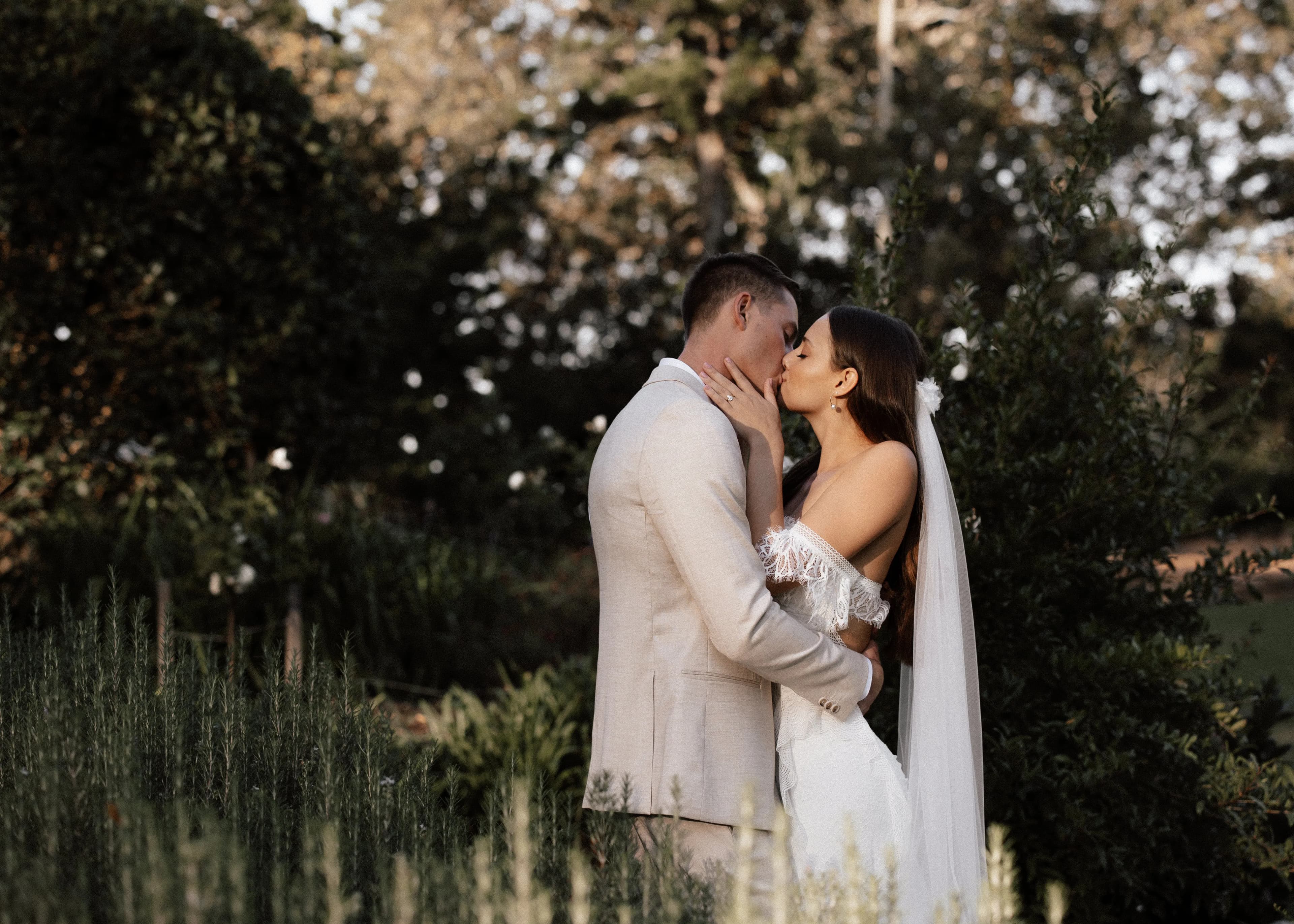 A bride and groom share a romantic kiss outdoors, surrounded by lush greenery. The bride wears an off-the-shoulder white wedding dress and a long veil, while the groom is dressed in a beige suit. The background features trees and foliage in soft, warm lighting.