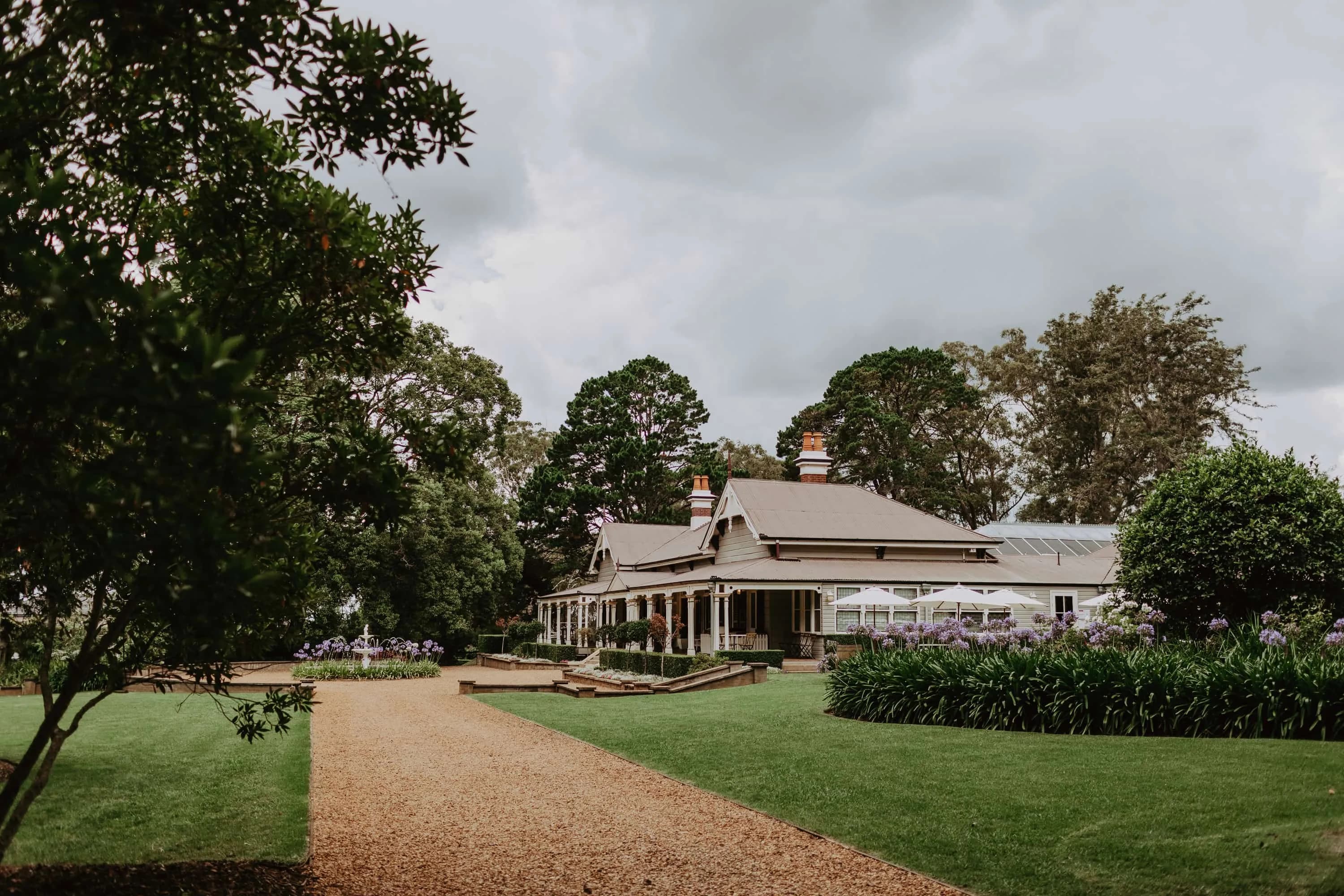 A large, historic wooden house with a wrap-around porch sits at the end of a gravel pathway, surrounded by manicured lawns and dense greenery. The sky above appears overcast, and purple flowers add color to the well-maintained garden.