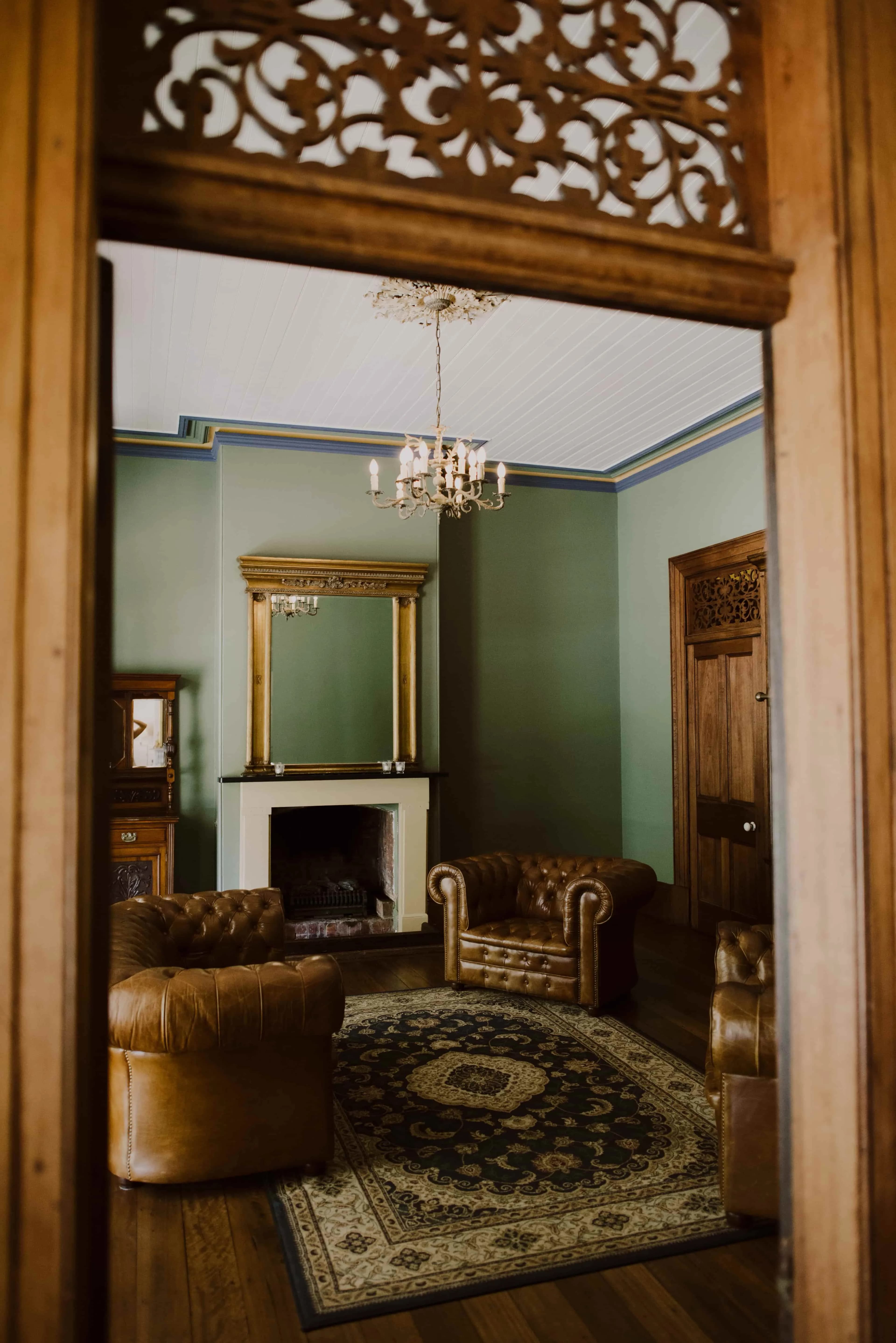 A cozy living room features green walls, a fireplace with a large mirror above it, and a vintage chandelier. The room has brown leather armchairs arranged around a patterned rug. Wooden doors and ornate woodwork add a classic, elegant touch to the space.