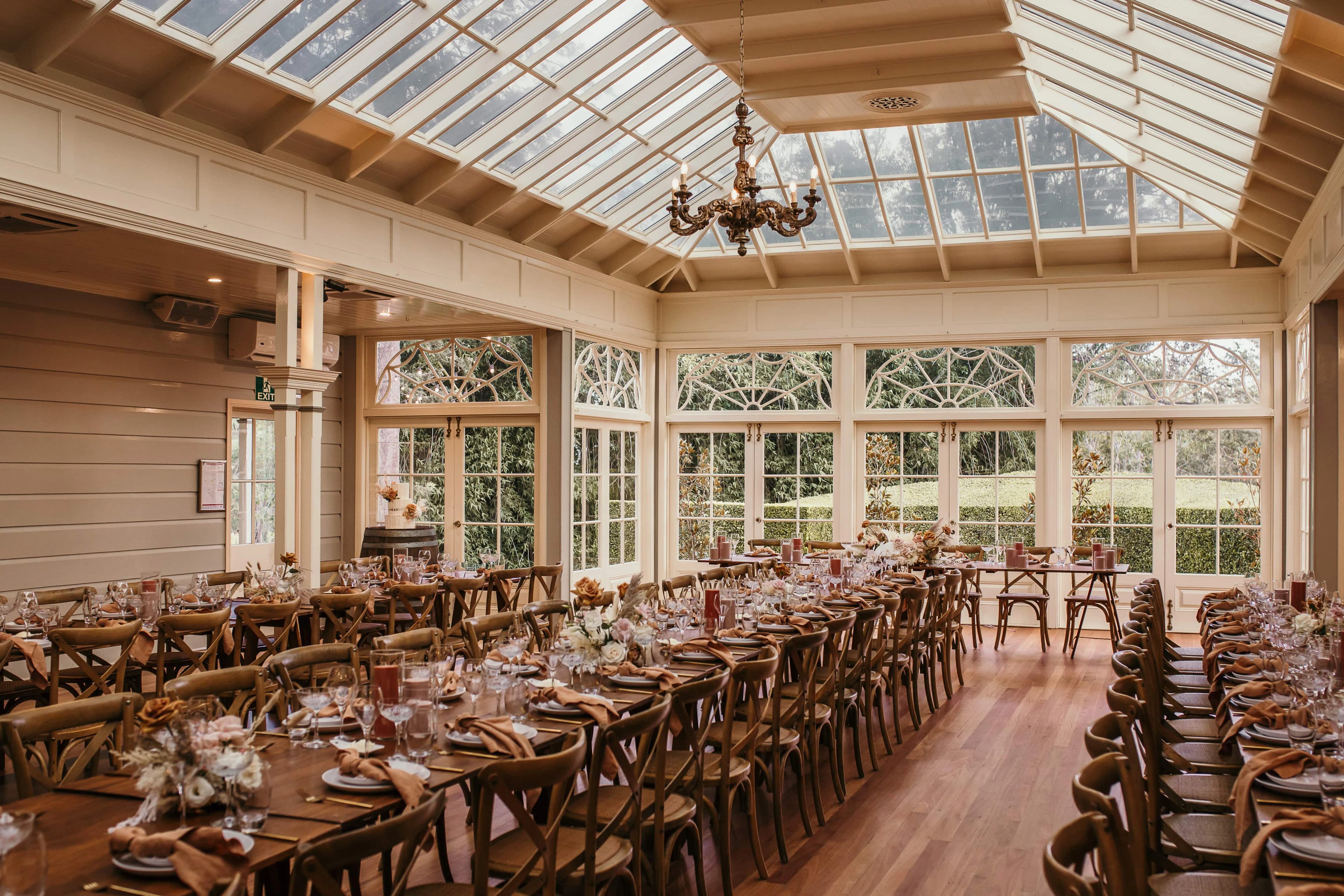 A spacious, elegantly decorated dining hall with large, paneled windows and a glass ceiling allowing natural light to flood in. Long wooden tables are set with fine tableware, floral centerpieces, and neatly folded napkins. A chandelier hangs from the ceiling.
