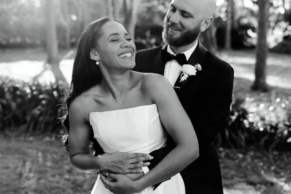 A joyful couple on their wedding day. The groom, dressed in a black tuxedo, stands behind the bride, who wears a strapless white gown. The bride is smiling and laughing, her eyes closed, as they share a tender embrace outdoors with trees in the background.