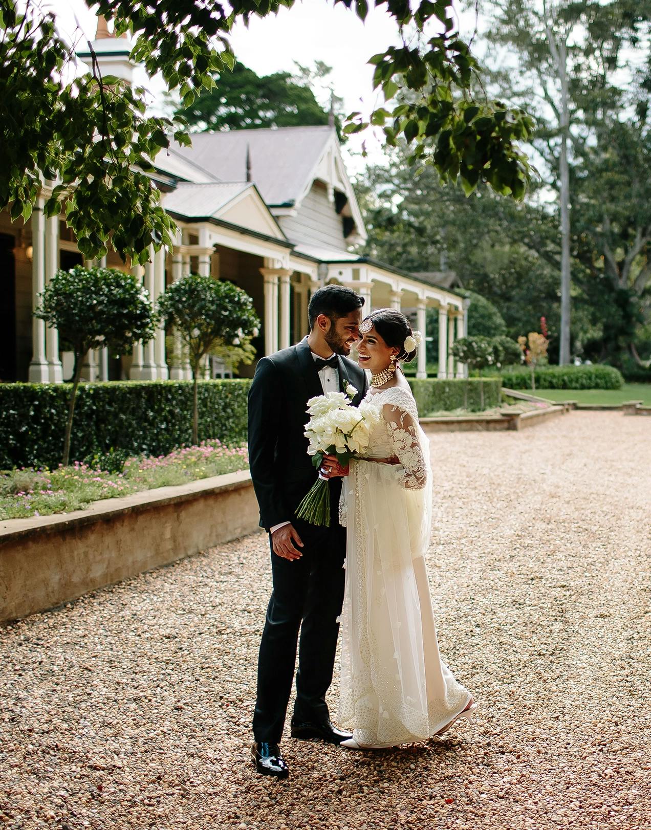 A bride and groom stand close together on a gravel path in front of a charming building, surrounded by greenery. The bride, in a white lace gown, holds a bouquet of white flowers, while the groom, in a black suit, leans in affectionately.