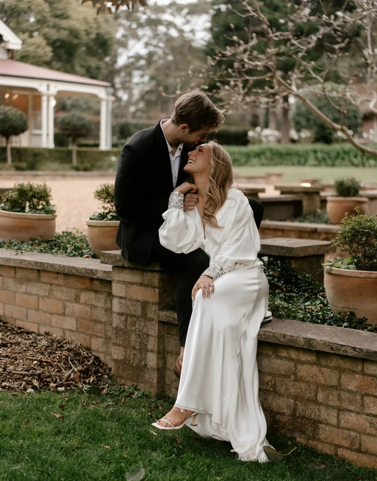 A couple smiles affectionately at each other in an outdoor garden setting. The man, dressed in a black suit, sits on a low brick wall while the woman, in a flowing white dress, stands close to him, holding his hand. Potted plants and trees surround them.