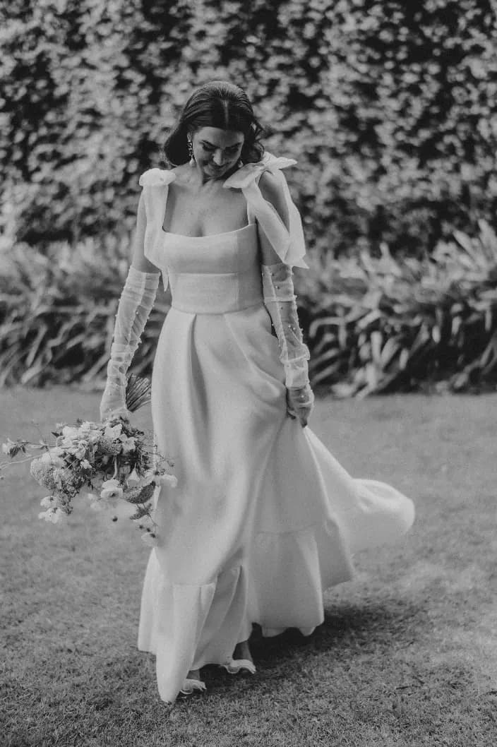 A woman in a flowing wedding dress walks through a garden. She holds a bouquet of flowers and is surrounded by greenery. The image is in black and white, highlighting the elegant design of her gown and the serene setting.