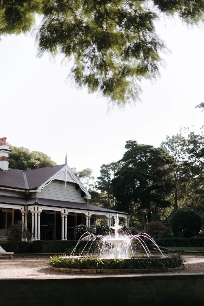 A classic wooden house with a peaked roof sits surrounded by lush greenery. In the foreground, a fountain sprays water into the air, and large tree branches partially frame the scene above.