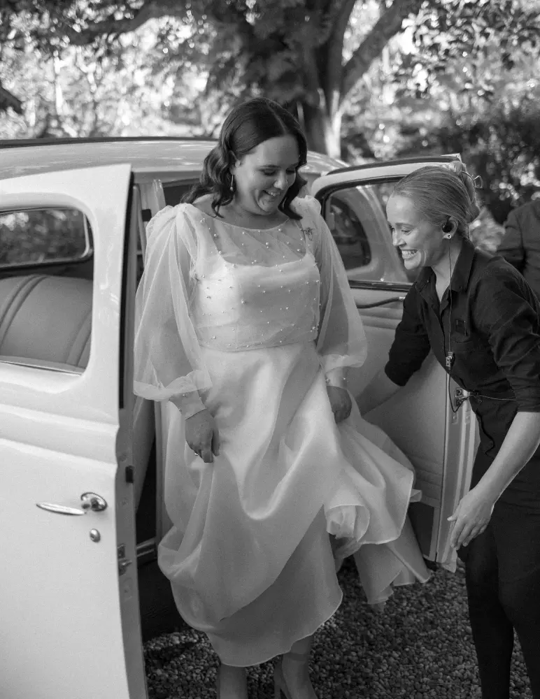 A bride in a flowing dress is stepping out of a classic white car. She is smiling and assisted by another woman. The scene is outdoors, surrounded by trees. The photo is in black and white.