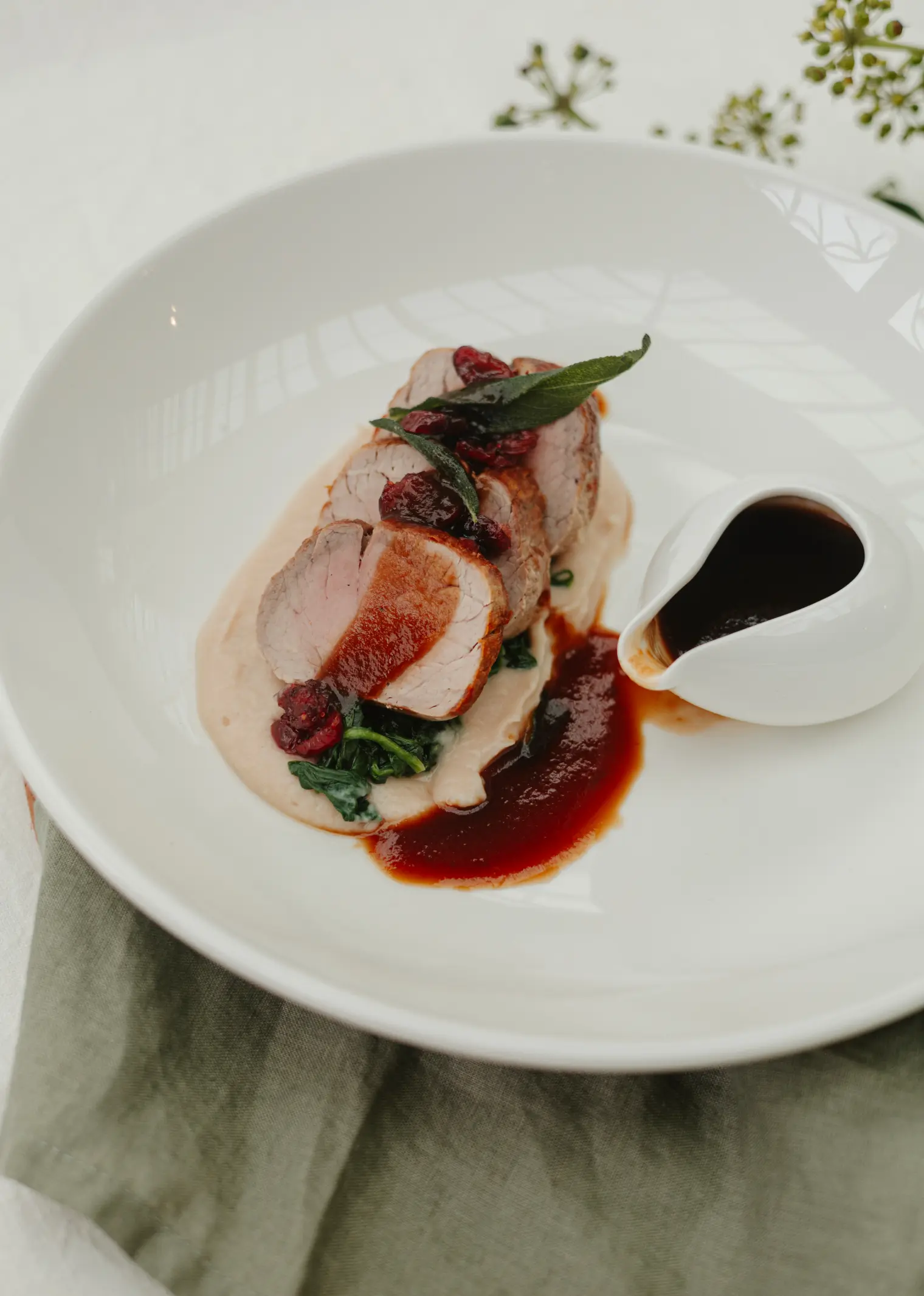 A gourmet dish featuring two slices of seared meat atop a bed of wilted greens and creamy puree, garnished with a leaf. A small white pitcher of dark sauce is on the side. The plate rests on a white tablecloth with a green napkin.