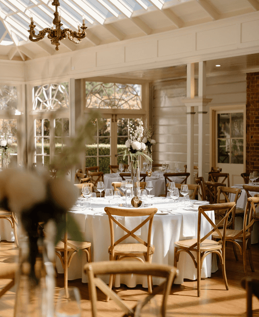 Elegant indoor wedding reception setup with round tables covered in white tablecloths. Each table is adorned with floral centerpieces and surrounded by wooden chairs. Sunlight streams through large windows and a glass ceiling, illuminating the room.