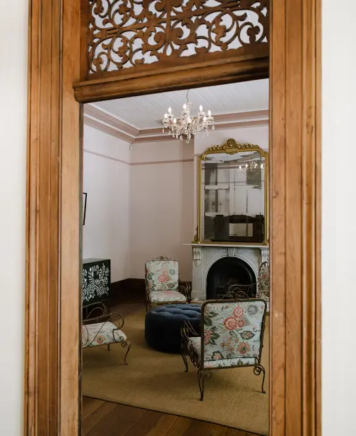 Elegant living room seen through a wooden doorway, featuring ornate furniture with floral patterns, a decorative fireplace with a large mirror above, and a sparkling chandelier hanging from the ceiling. Warm, inviting atmosphere.