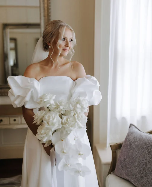 A bride in a white wedding dress stands by a window, holding a bouquet of white flowers. The dress features puffed sleeves, and her hair is styled up with loose curls. She gazes to the side, with soft natural light illuminating the scene.