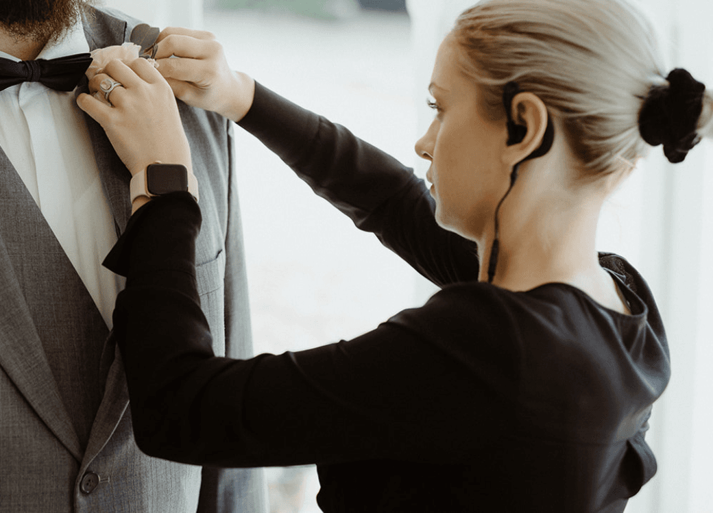 A woman adjusts a man's bow tie on his suit. She has her hair in a bun, wears a smartwatch, and has an earpiece in. The man’s suit is gray with a white shirt.