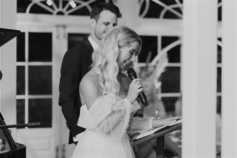 A black and white photo shows a woman in a formal off-shoulder dress speaking into a microphone while reading from a stand. A man in a suit stands nearby, slightly behind her, and smiles. The background features large windows with decorative framing.