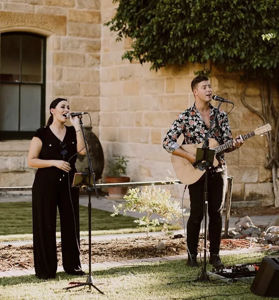 A woman and a man perform outdoors, both singing into microphones. The woman stands on the left, wearing a black outfit, while the man on the right plays a guitar and wears a floral shirt. They stand in front of a building with stone walls and a large window.