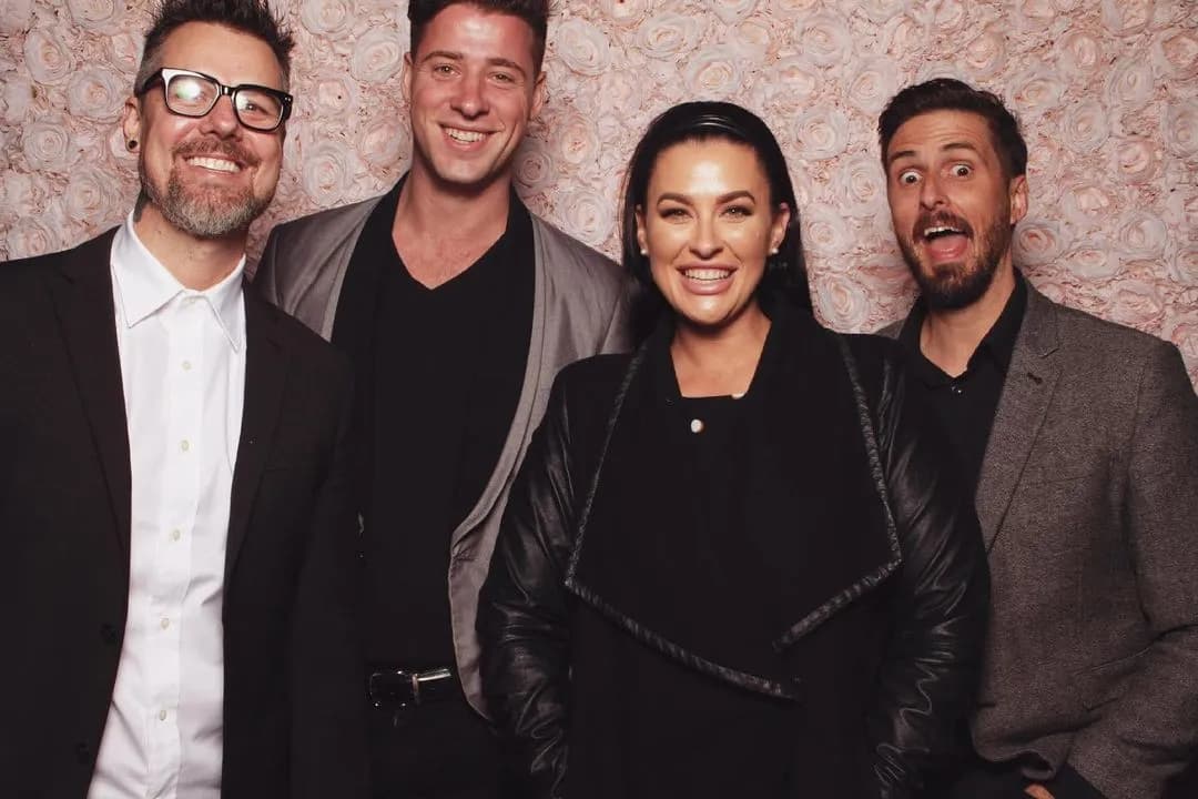 Four people are posing and smiling in front of a flower-patterned backdrop. The group consists of three men and one woman, all dressed in semi-formal attire. One of the men is wearing glasses. The atmosphere appears to be cheerful and friendly.