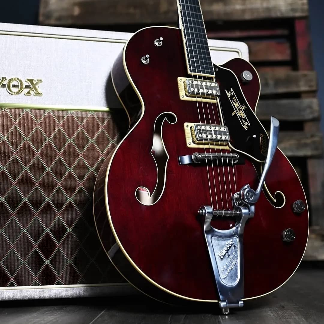 A close-up of a vintage red electric guitar leaning against a VOX amplifier. The guitar, featuring gold hardware and white trim, has f-holes and a Bigsby vibrato tailpiece. The amplifier is beige with a diamond-patterned cloth front and "VOX" branding.