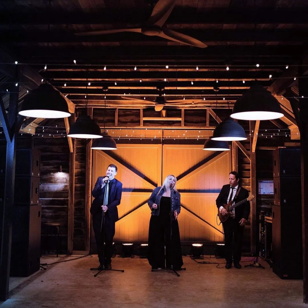A band performing in a rustic venue with wooden walls and string lights. The group comprises two vocalists, a man and a woman, on microphones, and a guitarist to the right. They are under industrial-style pendant lights, creating a warm, intimate atmosphere.