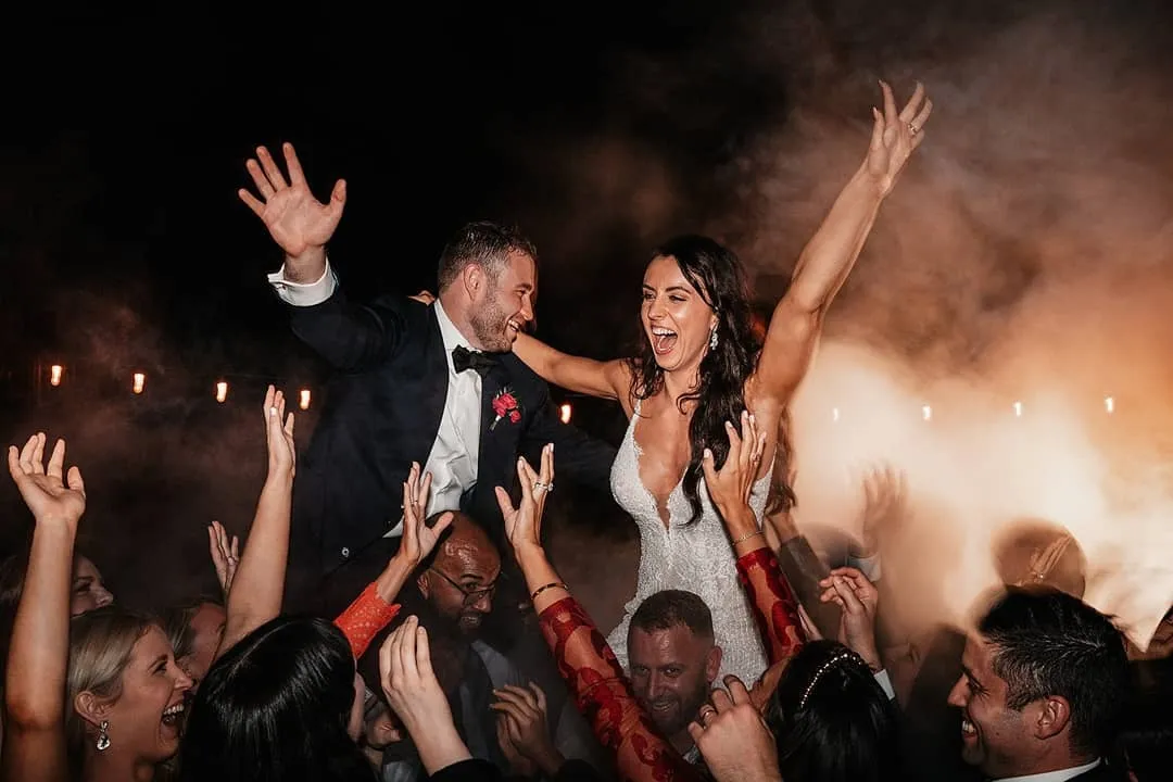 A joyous bride and groom in wedding attire are being lifted on chairs by a group of happy guests. The bride is wearing a white gown, and the groom is in a black tuxedo. Both have their arms raised in celebration. Fairy lights glow softly in the background amidst light smoke.