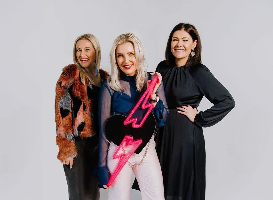 Three smiling women pose together against a plain background. The woman on the left wears a multicolored fur jacket, the woman in the center holds a black guitar with pink lightning bolts, and the woman on the right wears a black dress.