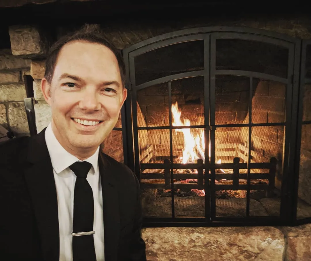A man in a black suit and tie smiles at the camera while sitting in front of a lit fireplace with a stone surround and metal grill.