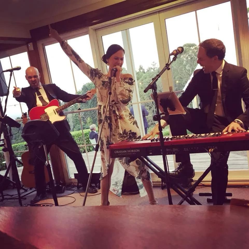 A woman in a floral dress sings passionately into a microphone while two men accompany her, one playing a red electric guitar and the other playing a keyboard. The scene appears to be in a cozy indoor setting with large windows showing a garden outside.