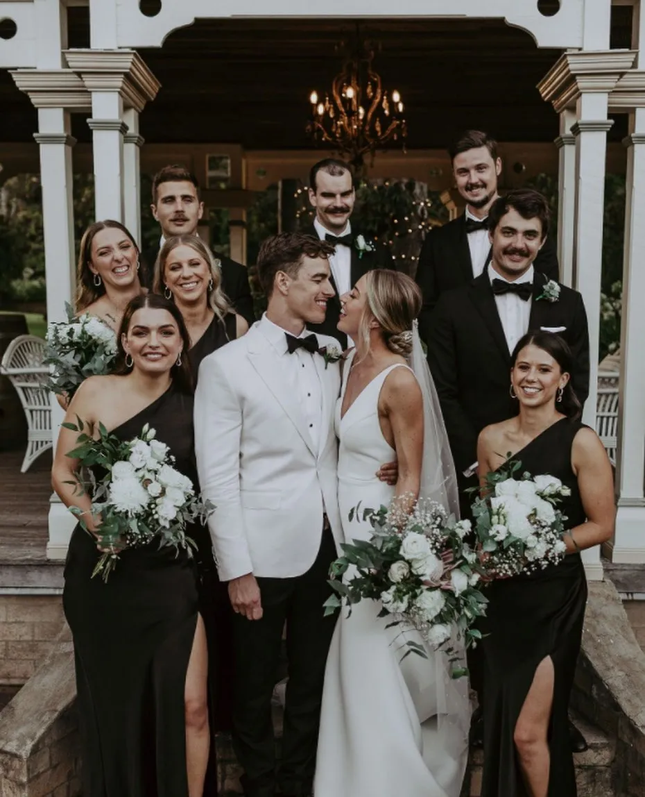 A bride and groom stand smiling at each other in front of a porch, surrounded by their wedding party. The bridesmaids are wearing black dresses, and the groomsmen are in black tuxedos with bow ties. Everyone is holding bouquets and looking towards the couple.