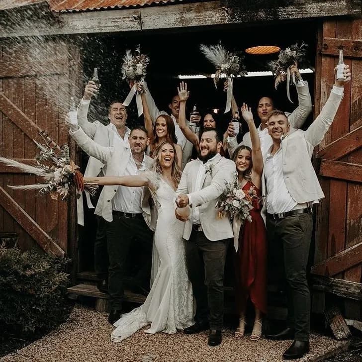 A wedding party celebrates outside a rustic wooden barn. The bride and groom stand in the center, holding hands and smiling, surrounded by bridesmaids and groomsmen. Everyone is cheering with raised arms and drinks, and one person is spraying a bottle of champagne.