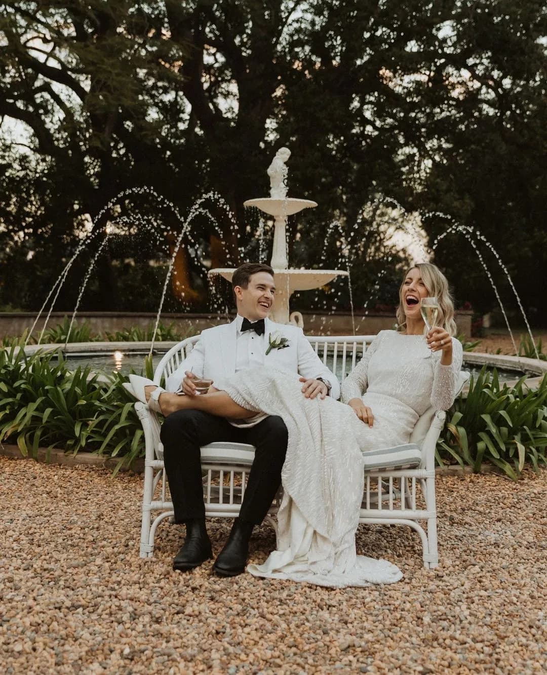 A couple, both dressed in white formal attire, sits on a white bench in front of a fountain. The man holds a drink while the woman, also holding a drink, laughs joyfully. They appear to be celebrating, surrounded by greenery and gravel ground.