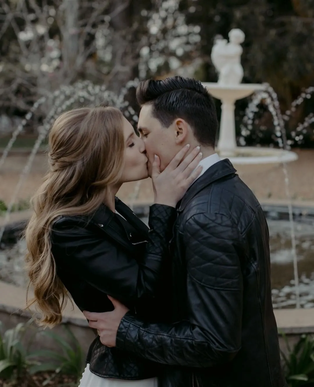 A couple dressed in black leather jackets shares a kiss in front of a fountain. The woman has long, wavy hair, and the man has short, dark hair. The background features a statue and a blurred, outdoor setting with trees.