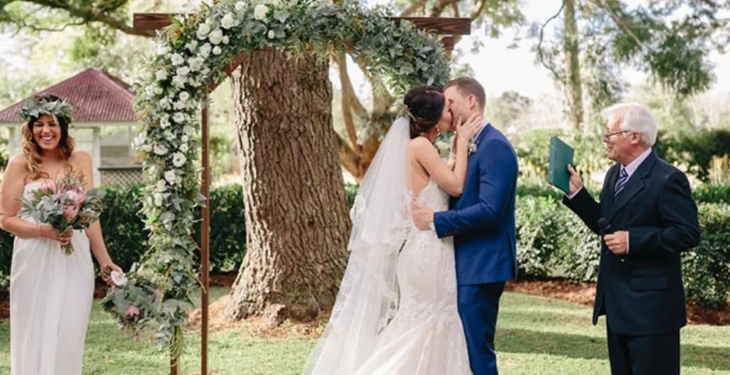 A bride and groom are kissing under a floral arch during an outdoor wedding ceremony. The bride, in a white dress, holds the groom's face. The groom is dressed in a blue suit. A joyful bridesmaid stands to the side, and an officiant holds a book on the other side.