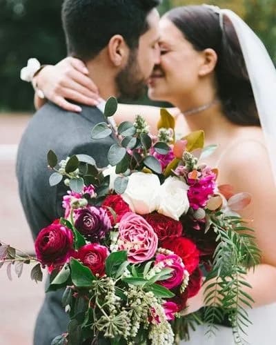A bride and groom share a tender kiss on their wedding day. The bride, in a white dress and veil, holds a large, vibrant bouquet of pink, red, and white flowers with greenery. The groom, in a gray suit, embraces her, and both appear happy and in love.