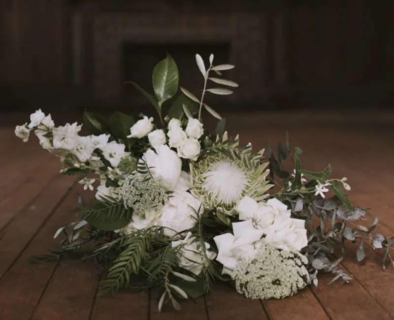 A floral arrangement featuring white flowers and green foliage rests on a wooden floor. The bouquet includes a variety of blooms, such as roses and chrysanthemums, complemented by leafy greens and sprigs of eucalyptus. The background is dimly lit with a wooden surface.