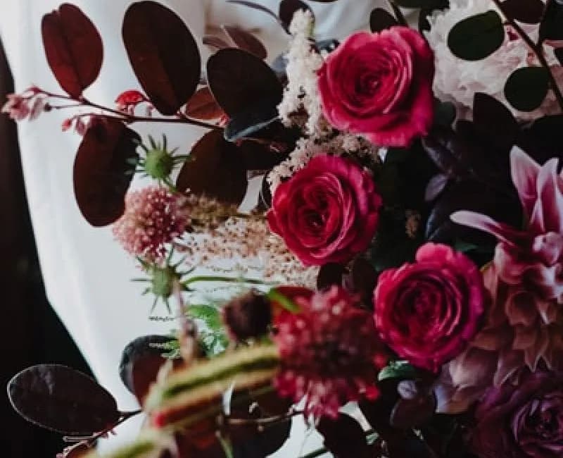 Close-up of a vibrant bouquet featuring red roses, deep purple foliage, and various other flowers. The background is softly blurred, possibly showing a person holding the bouquet. The mix of colors and textures creates a lush, romantic appearance.