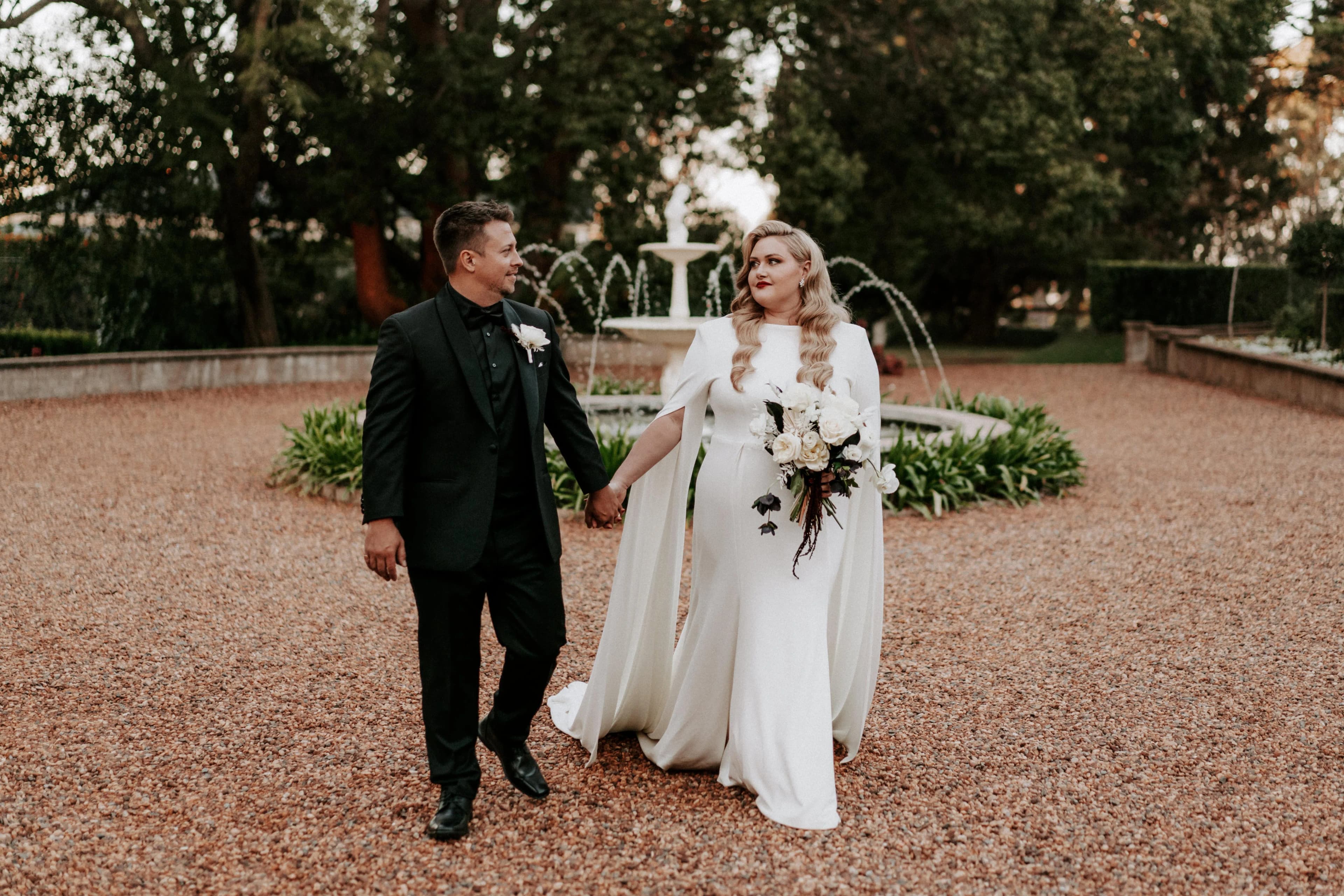 A couple walks hand-in-hand in front of a fountain surrounded by greenery. The woman is in a white gown holding a bouquet, while the man wears a black suit. Both look at each other lovingly. The setting appears to be an outdoor garden.