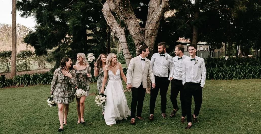 A bride and groom hold hands, walking with a joyful wedding party in a lush, green outdoor setting. The wedding party consists of three bridesmaids in floral dresses and three groomsmen in white shirts and black pants. Everyone is smiling and chatting under the trees.