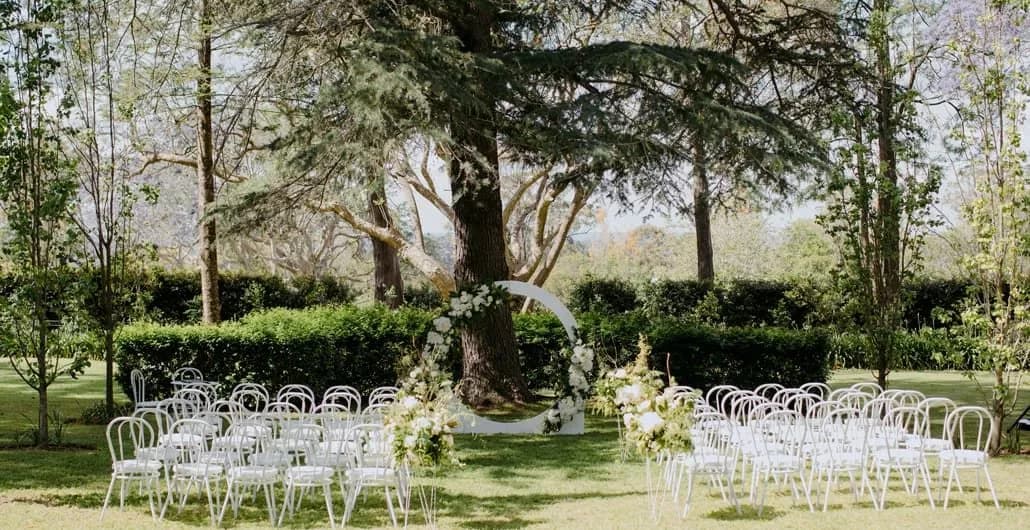 An outdoor wedding setup featuring rows of white chairs arranged on green grass, facing a large tree adorned with white flowers and a circular floral arch set at its base. The backdrop includes lush green hedges and tall trees.