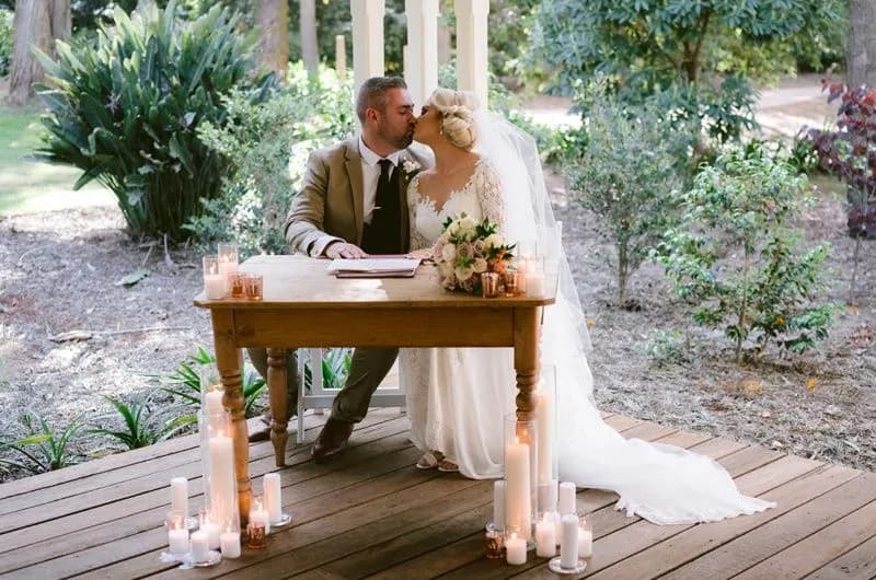 A bride and groom sit at a wooden table outdoors in a garden. The groom, in a beige suit and tie, kisses the bride, who wears a white lace wedding dress and veil. The table is adorned with flowers and surrounded by lit candles. The background is lush with greenery.