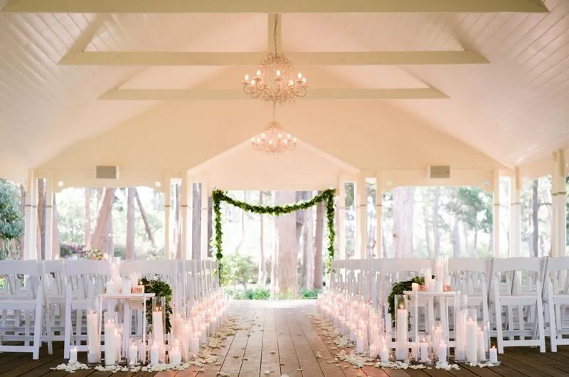 An elegantly decorated indoor wedding chapel with white wooden chairs arranged in rows. The aisle is lined with candles and greenery. Flower petals are scattered along the wooden floor. A garland of greenery adorns the front, framing large windows with a view of trees.