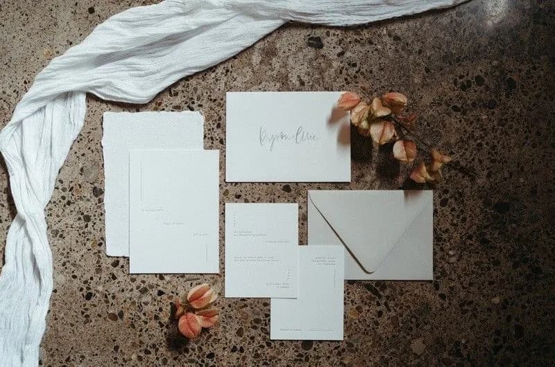 A flat lay of wedding stationery and decor on a speckled surface. The set includes invitation cards, an envelope, and menu cards arranged aesthetically. Surrounding them are light pink flower buds and a flowy white fabric. The invitation reads "Reyna & Allen".