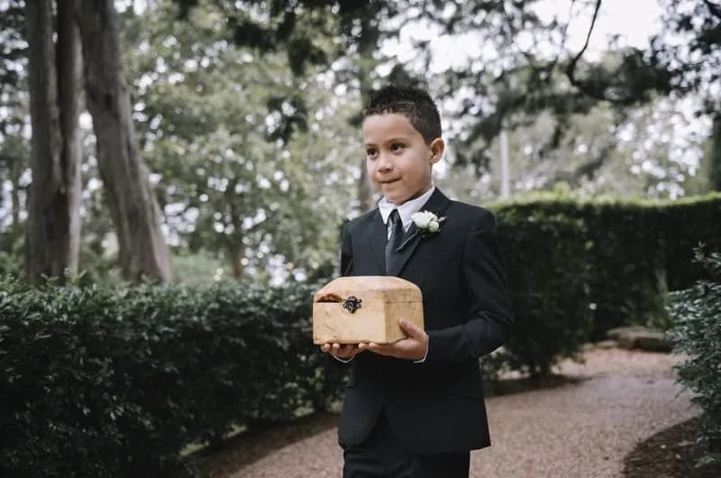 A young boy in a black suit and tie, with a white boutonnière on his lapel, holds a wooden box while walking along a garden path lined with greenery and trees. The setting appears to be outdoors, possibly a wedding or formal event.