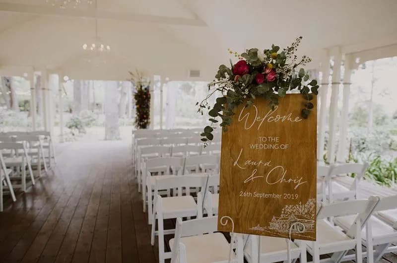 A wedding venue with white chairs arranged in rows on a wooden floor. At the entrance, there's a wooden sign adorned with greenery and red flowers that reads, "Welcome to the wedding of Laura & Chris, 26th September 2019." The venue is airy with lots of natural light.