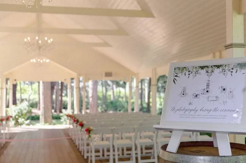 An outdoor wedding venue features a white canopy and rows of white chairs adorned with red flowers on the aisle ends. A framed sign on an easel in the foreground reads, "No photography during the ceremony," with illustrated cameras and greenery accents.