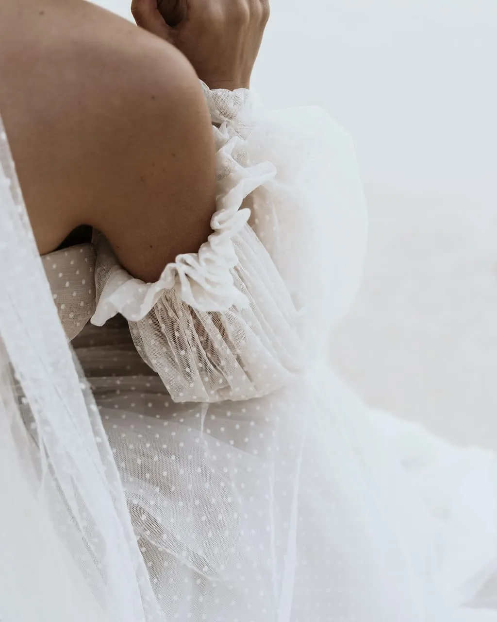 Close-up of a person wearing a sheer, white, polka-dot dress with gathered, puffed sleeves. The person's bent arm and shoulder are visible, and the delicate fabric drapes softly, creating an elegant and ethereal look. The background is blurred and neutral in tone.