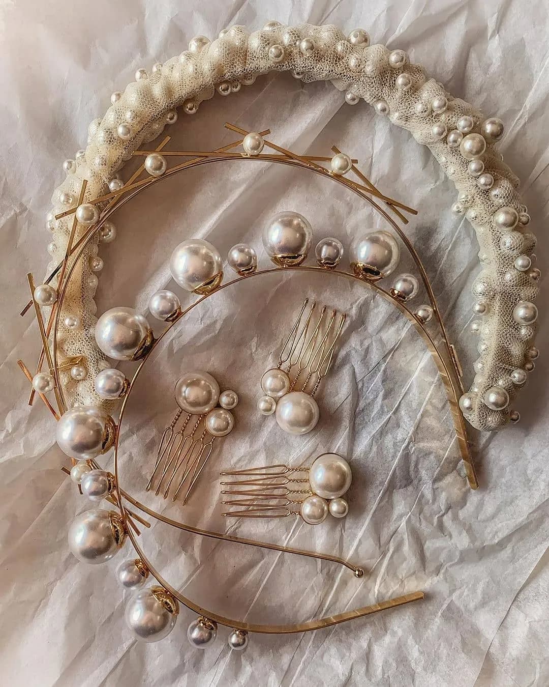 A collection of bridal hair accessories, including a pearl-embellished headband, two gold hairpieces adorned with various sizes of pearls, and three pearl hairpins, all arranged on crinkled white paper.