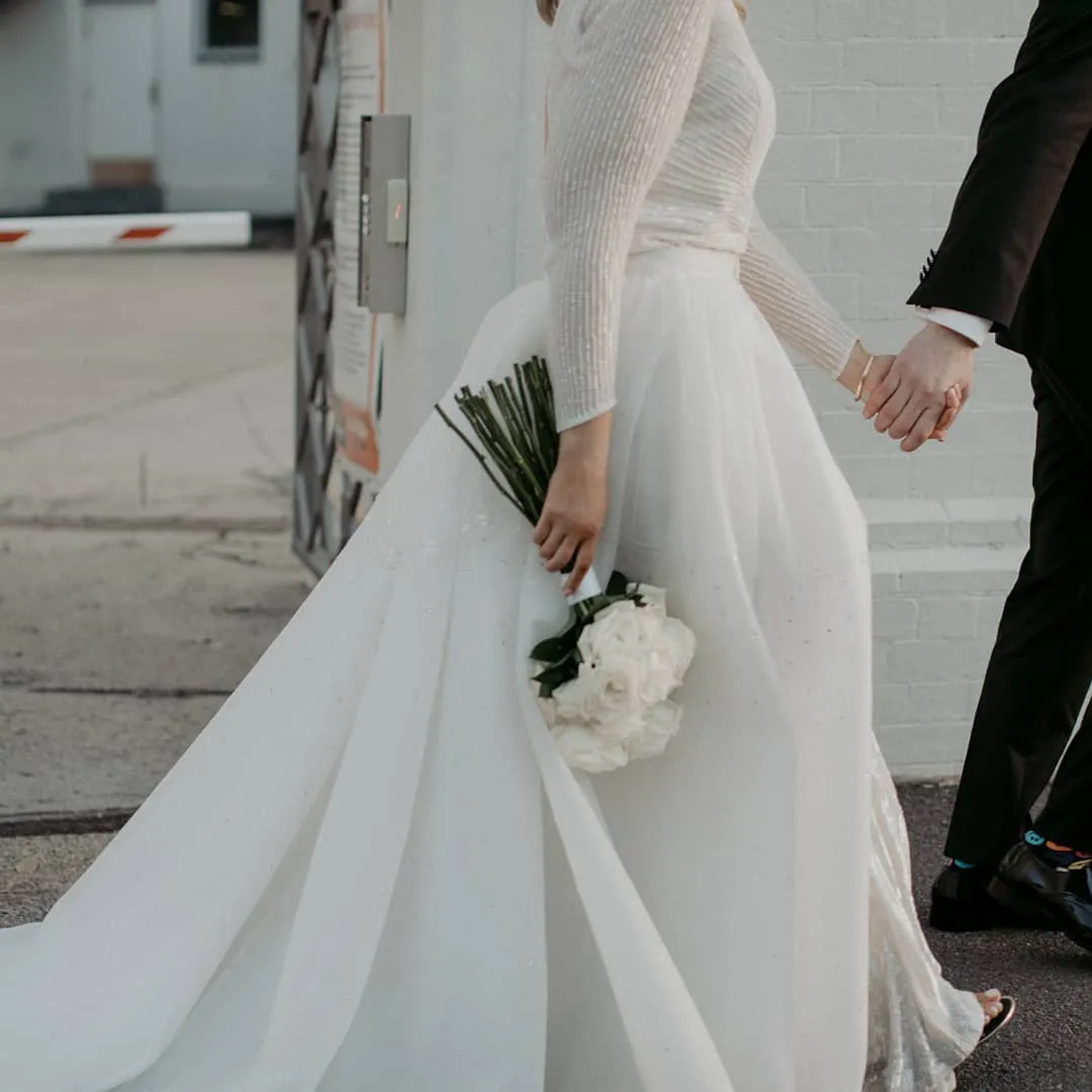 A bride in a white wedding gown holds a bouquet of white flowers, walking hand-in-hand with someone in a black suit. The image captures a side view of the couple as they walk outdoors near a building.